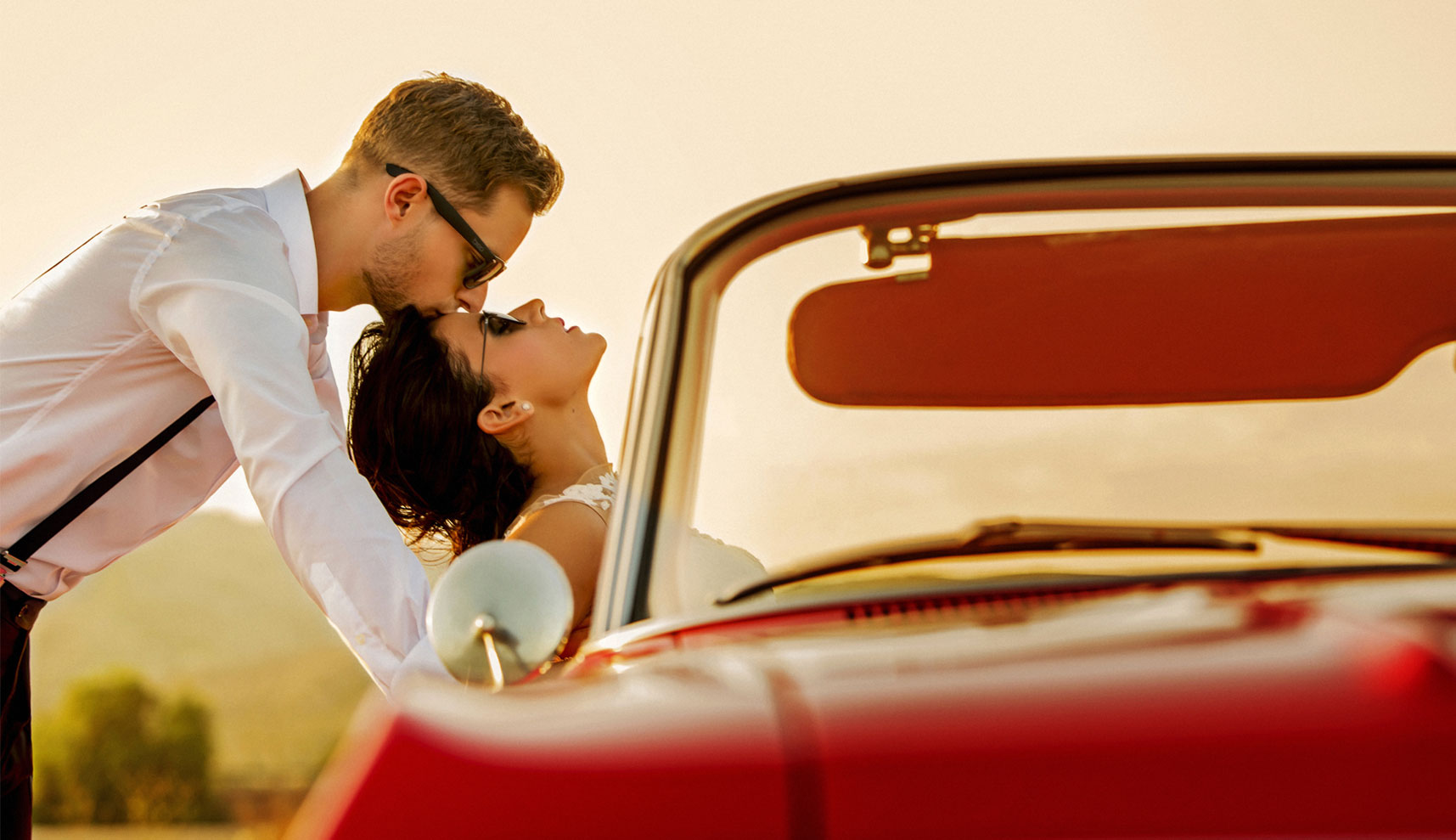 El novio dándole un beso en la frente dónde ella esta apoyada en el coche.